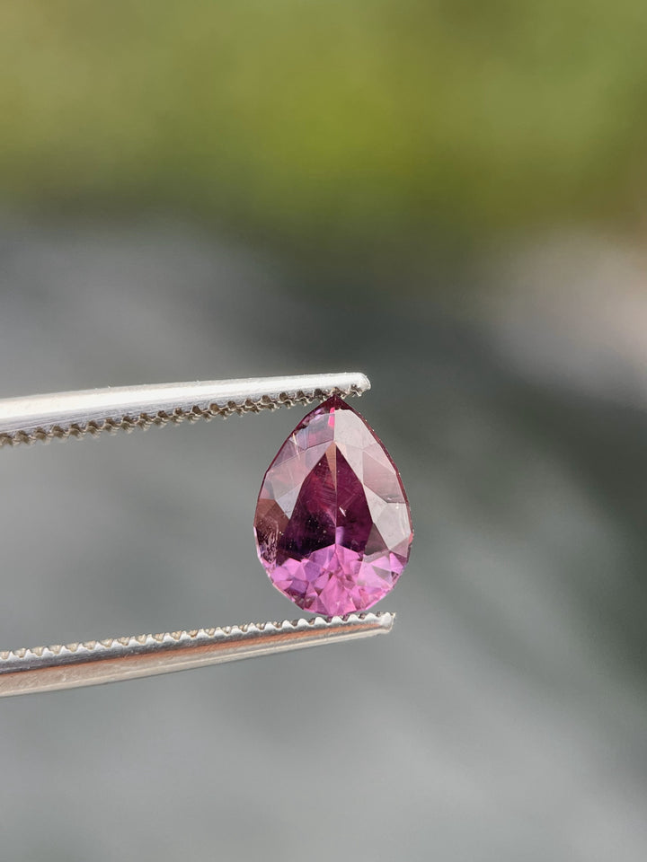A pear-shaped pinkish-purple sapphire weighing 1.45 carats held by metal tweezers against a blurred green background.