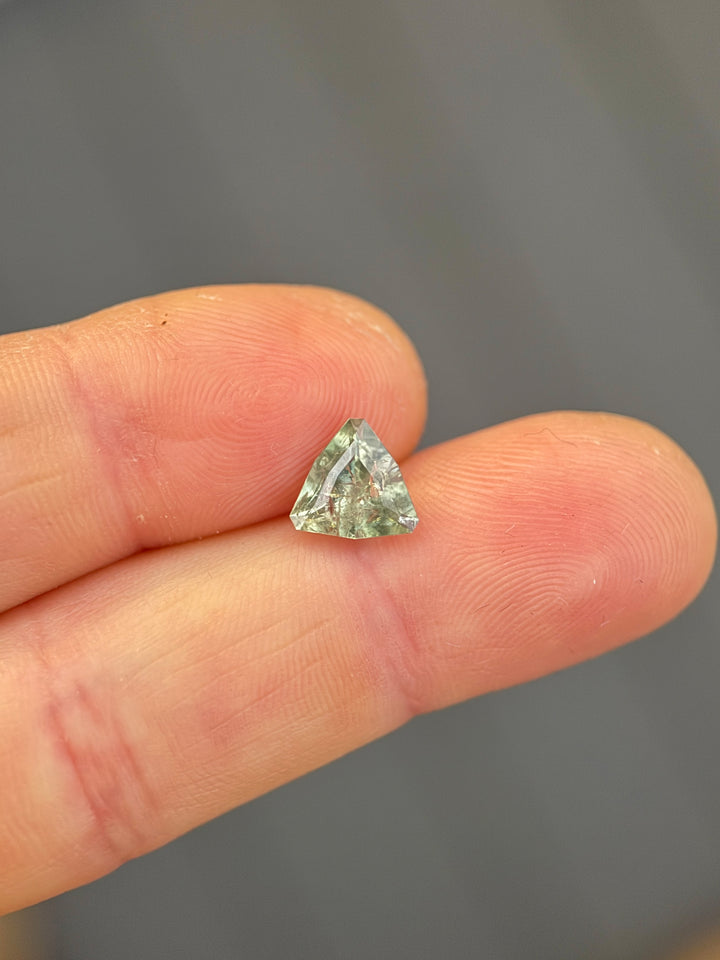 2.16 carat Montana sapphire in a custom geometric trillion cut resting on fingertips, showing yellow, teal, green, and sage color zoning with natural inclusions visible under macro lighting.