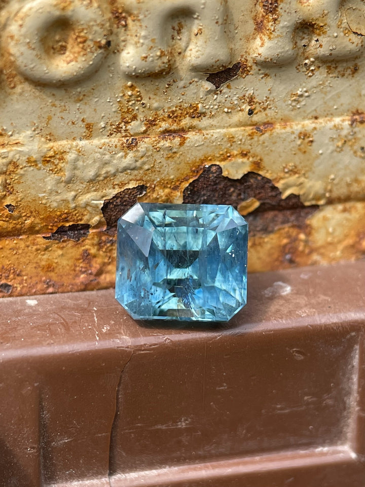 Asscher cut sapphire with teal, blue, and green hues weighing 2.50 carats, placed on a rustic metal background, mined from the El Dorado Bar Deposit in Montana.