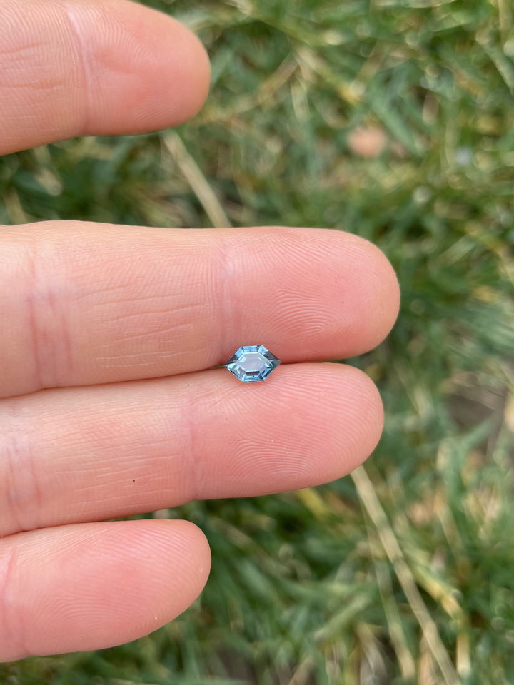 A 0.82-carat teal-blue Montana sapphire in a hexagonal open culet cut, resting on a fingertip. The stone displays precision faceting with a vibrant teal hue.