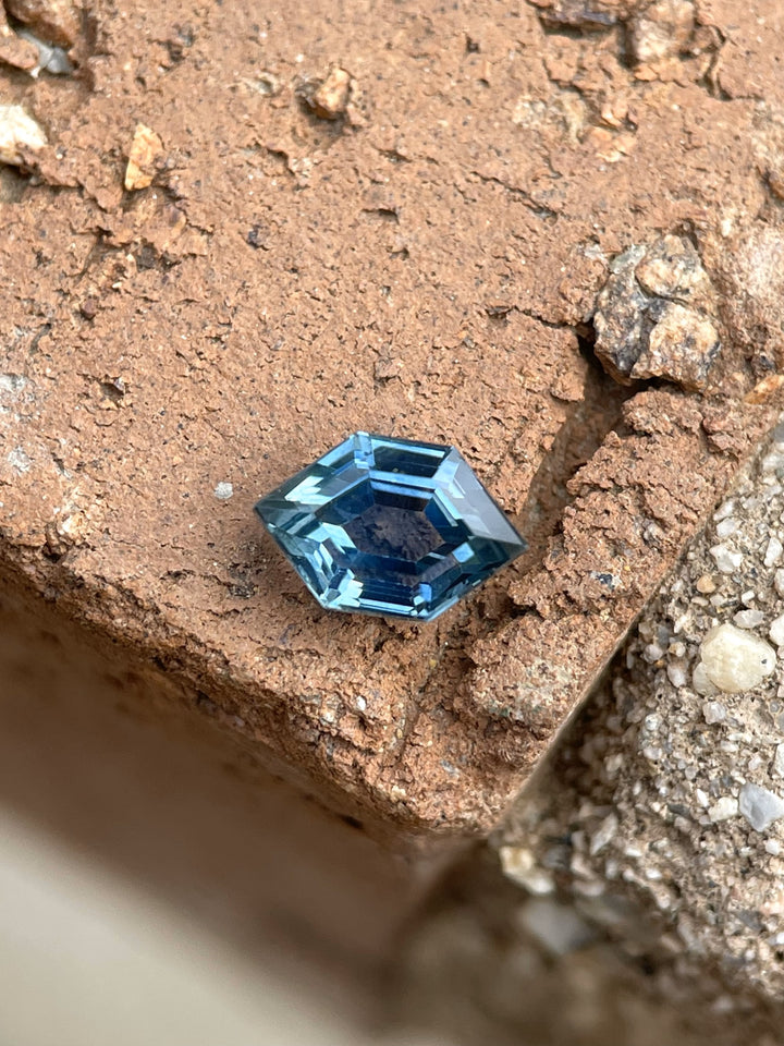A 0.82-carat teal-blue Montana sapphire with a hexagonal open culet cut, showcased on a rustic brick surface. The intricate facets highlight its teal and blue color tones.