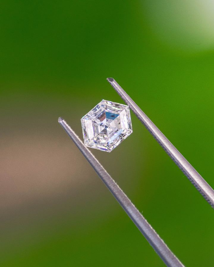 Hexagonal step cut diamond held by precision gem tweezers against a blurred green background. This natural 1.01 carat G color I2 clarity diamond from Gahcho Kué, Canada, showcases crisp faceting and mirror-like reflections.