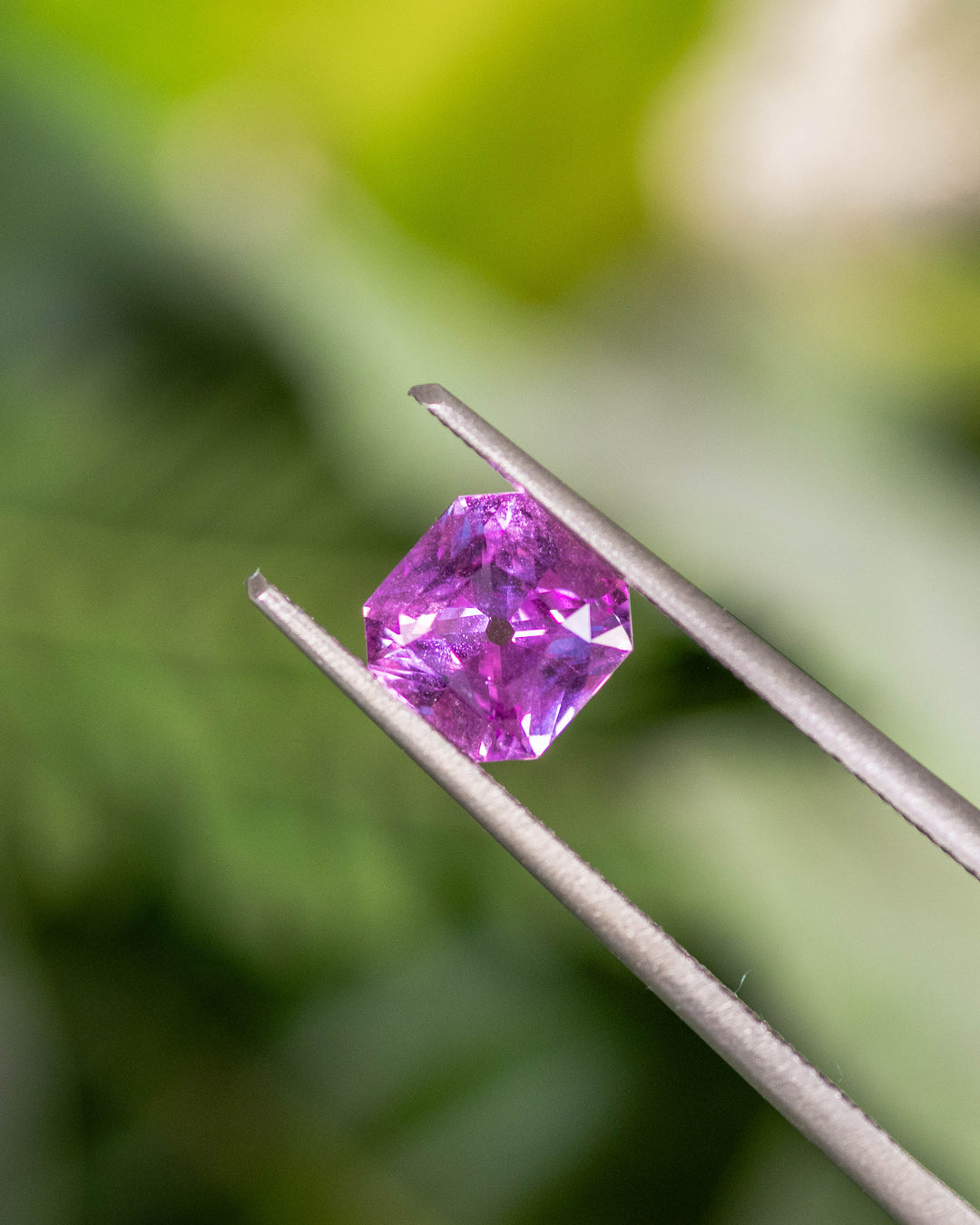 Close-up image of 1.03CT pink-purple sapphire held in gemstone tweezers under natural light, displaying sharp geometric facets and luster of the square scissor cut, unheated sapphire from Sakaraha, Madagascar.