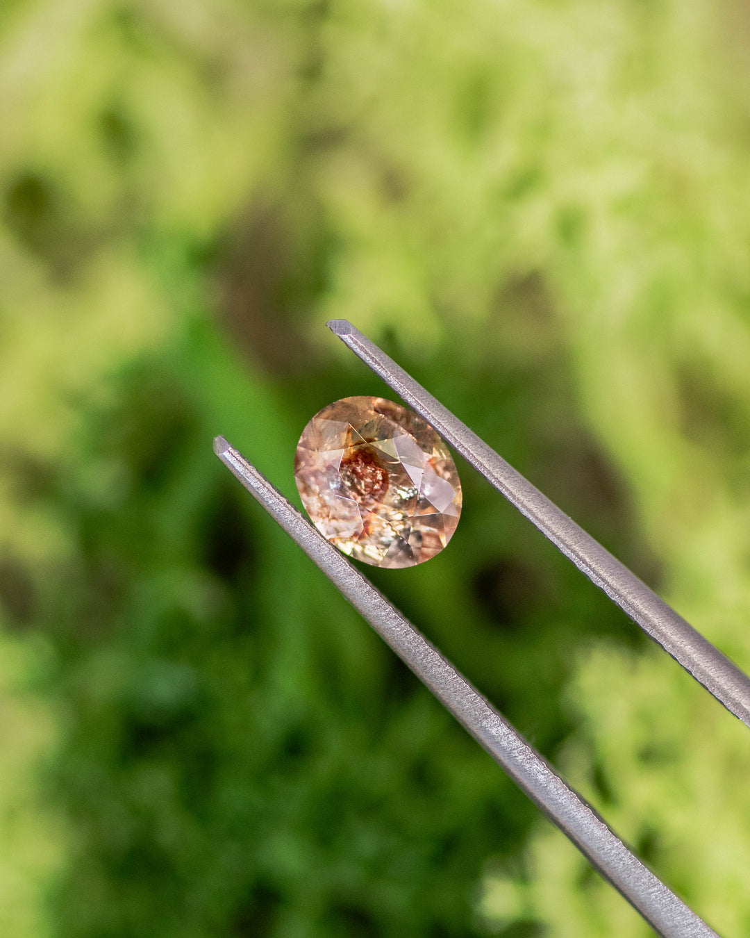 Close-up of 1.05CT orange peach red sapphire held in gemstone tweezers, revealing intricate internal inclusions and sparkle of custom oval cut, unheated sapphire from Umba River Valley, Tanga, Tanzania.