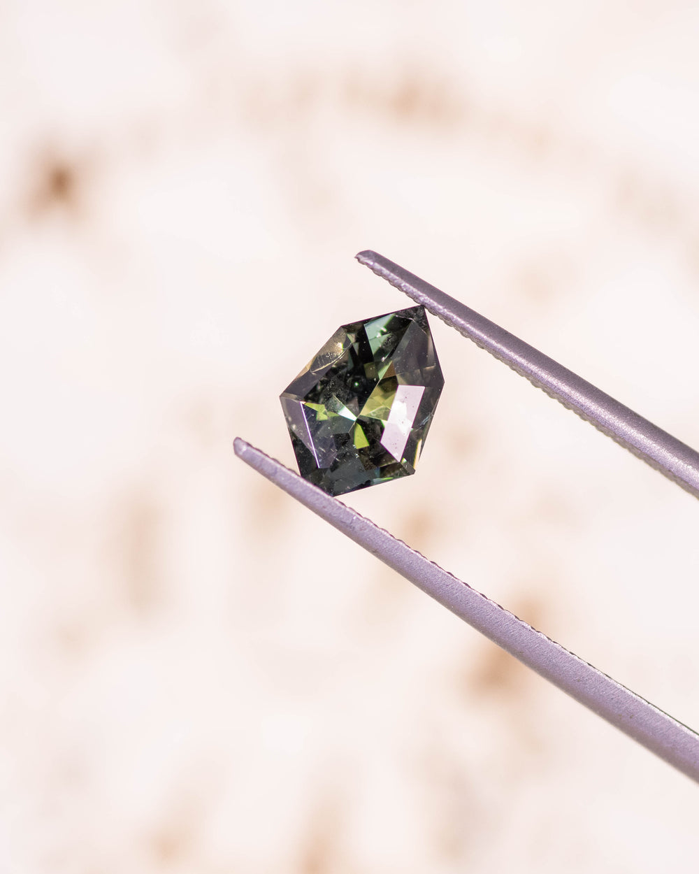 Macro image of 1.05CT yellow green blue tanzanite held in gemstone tweezers against a soft neutral background, highlighting precise facets and radiant flashes of color in this unheated geometric mix cut shield from Merelani Hills, Tanzania