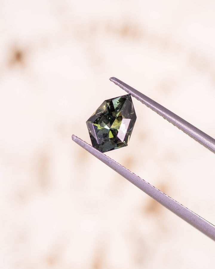 Macro image of 1.05CT yellow green blue tanzanite held in gemstone tweezers against a soft neutral background, highlighting precise facets and radiant flashes of color in this unheated geometric mix cut shield from Merelani Hills, Tanzania