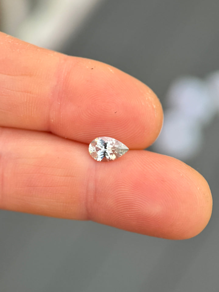 Precision pear cut Montana sapphire balanced on fingertips for scale, displaying a faint silver seafoam color, excellent light return, and natural untreated clarity in a 1.13 carat loose sapphire.