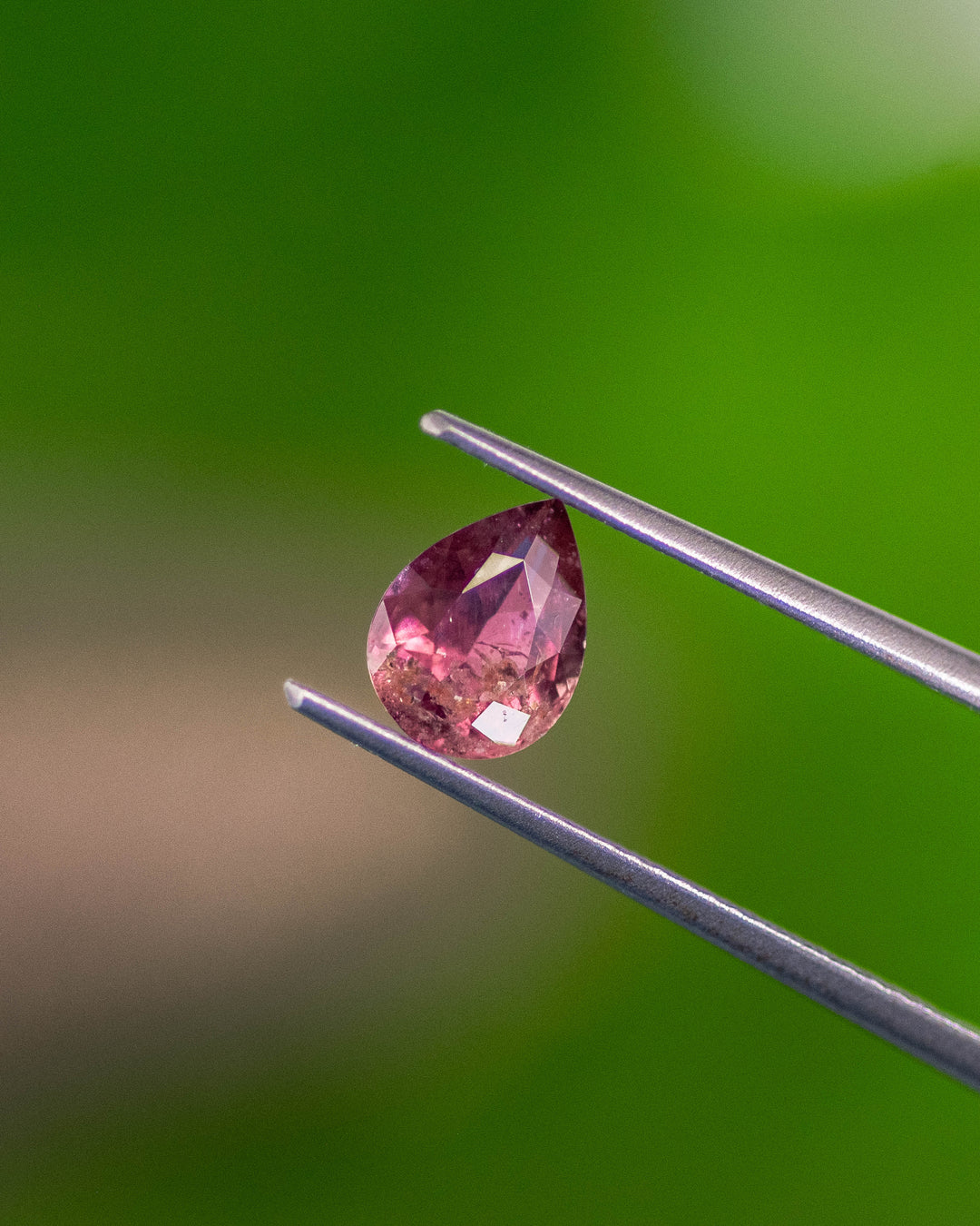 Close-up of 1.24CT orange pink red sapphire balanced on fingertip, showing the pear-shaped modified brilliant cut and internal inclusions under natural daylight, unheated gem from Umba River Valley, Tanzania.