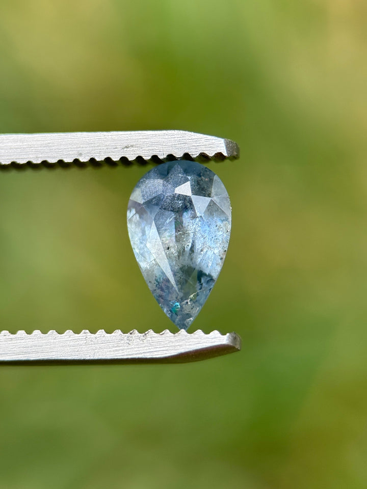 Close-up of 1.30CT pear mix cut sapphire held in tweezers under natural daylight, showing periwinkle blue tones and internal mineral textures, unheated gem from Umba River Valley, Tanzania.