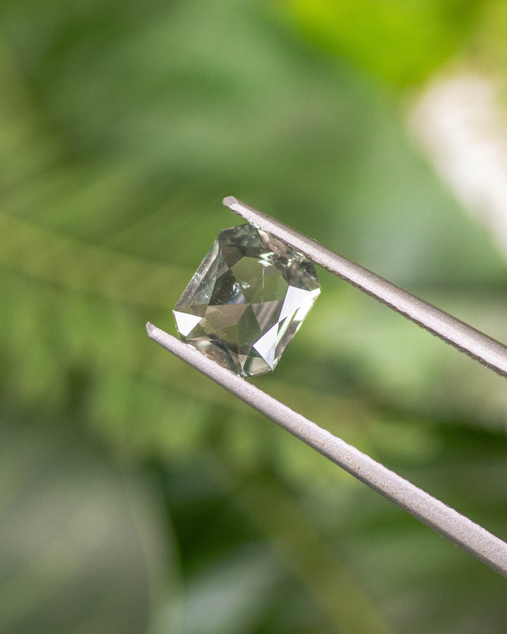1.31CT teal yellow green parti sapphire held in gemstone tweezers with lush greenery in the background, highlighting the facets and light reflection of its custom modified radiant cut, unheated sapphire from Umba River Valley, Tanzania.