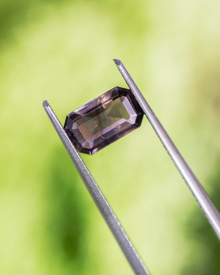 1.34CT purple violet eggplant sapphire held in gemstone tweezers with soft green natural background, highlighting facets and light reflection of its custom modified emerald cut, unheated sapphire from Umba River Valley, Tanzania.