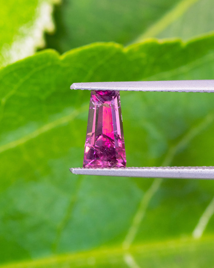 Close-up image of 1.39CT pink purple garnet held in gemstone tweezers under natural light, revealing deep color tones and geometric precision of the step cut tapered baguette, unheated garnet from Kenya.