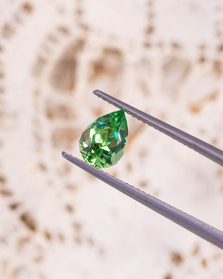 Macro image of 1.39CT tsavorite garnet held in gemstone tweezers against a soft beige background, highlighting its intense lime green sparkle and precise pear cut faceting, unheated gem from Kenya.