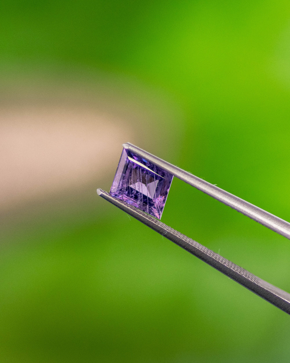 Macro photo of 1.40CT purple sapphire held in gemstone tweezers with a natural green background, highlighting its transparent luster, precise rhombus step cut, and unheated origin from Umba River Valley, Tanzania.