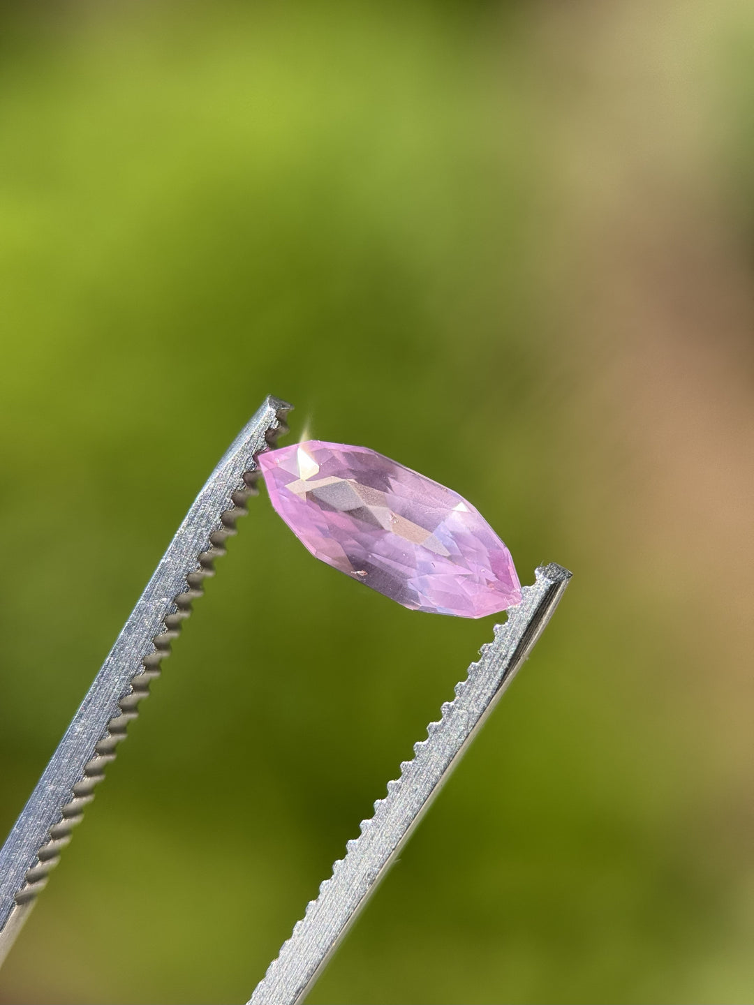 Close-up photo of 1.50CT bubblegum pink sapphire held in gemstone tweezers under natural light, showing luster and symmetry of the precision marquise mixed cut, unheated sapphire from Sakaraha, Madagascar.