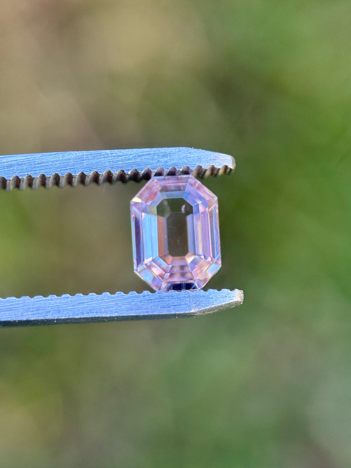 Macro image of 1.50CT blush pink sapphire held in gemstone tweezers against a blurred green background, highlighting the clean lines, transparency, and precision custom antique emerald cut of the unheated sapphire from Madagascar.