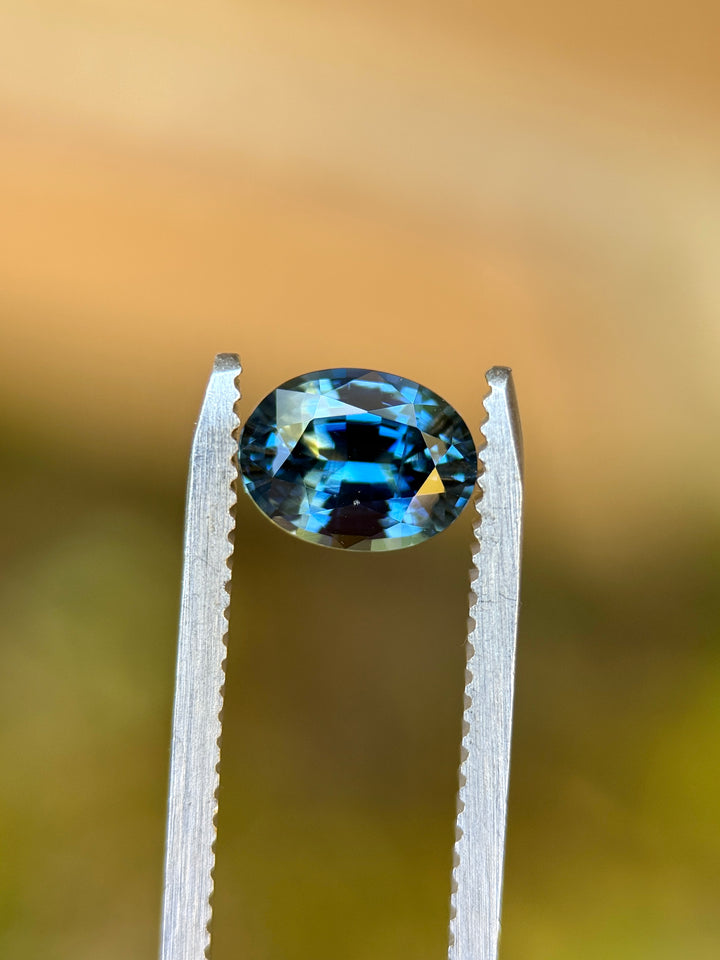Macro photo of 1.55CT yellow teal blue parti sapphire held in gemstone tweezers against a soft background, showcasing its vivid brilliance and precise oval mixed cut, unheated sapphire from Sakaraha, Madagascar.