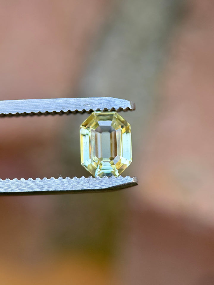 1.55 carat yellow sapphire held in gemstone tweezers under natural light, highlighting the crisp lines and geometric brilliance of the precision custom antidote emerald cut, untreated sapphire from Sakaraha, Madagascar.