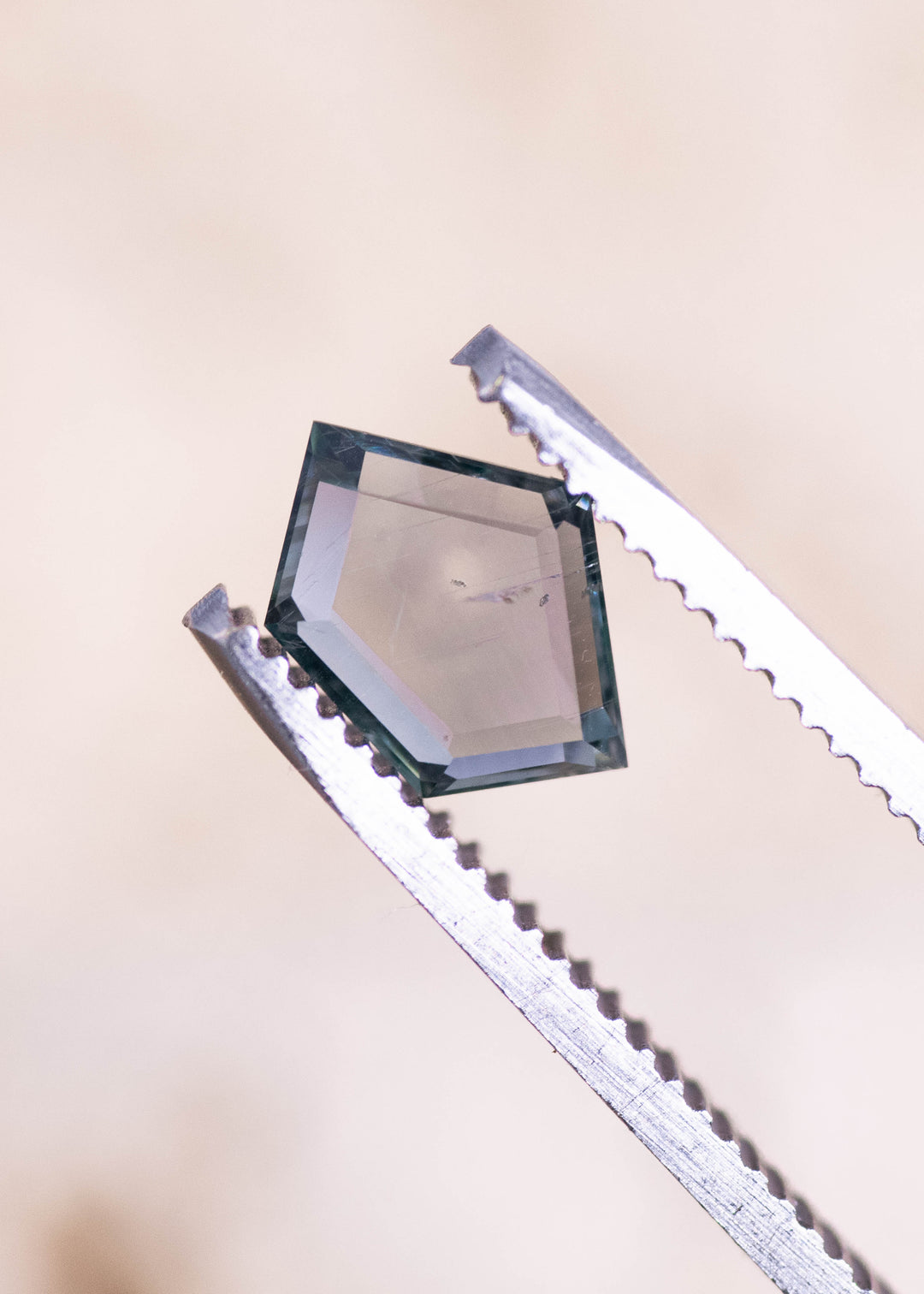 Side-angle view of a 1.57 carat Montana sapphire held in gem tweezers, highlighting the geometric pentagon portrait cut, crisp step facets, and teal blue seafoam body color.