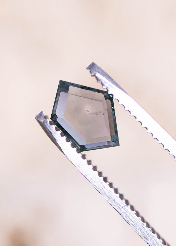 Side-angle view of a 1.57 carat Montana sapphire held in gem tweezers, highlighting the geometric pentagon portrait cut, crisp step facets, and teal blue seafoam body color.