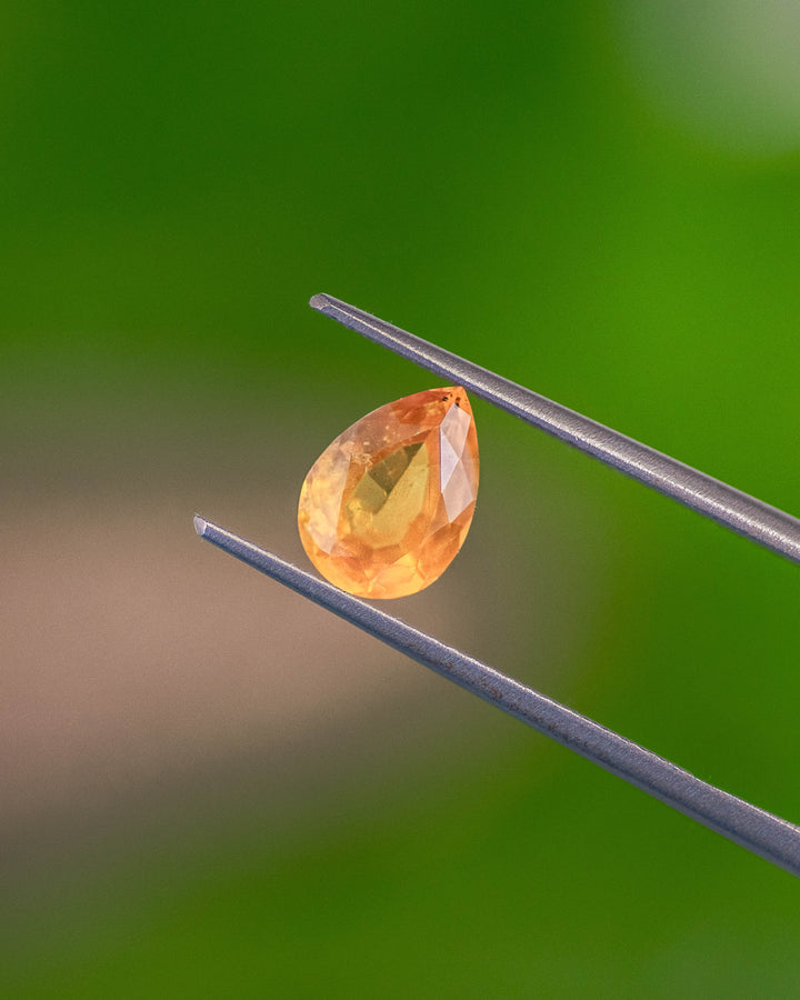 Macro image of 1.59CT red-orange and yellow sapphire held in gemstone tweezers against a green background, displaying its fiery tones and precision faceting of the modified brilliant pear cut, unheated gem from Tanzania.