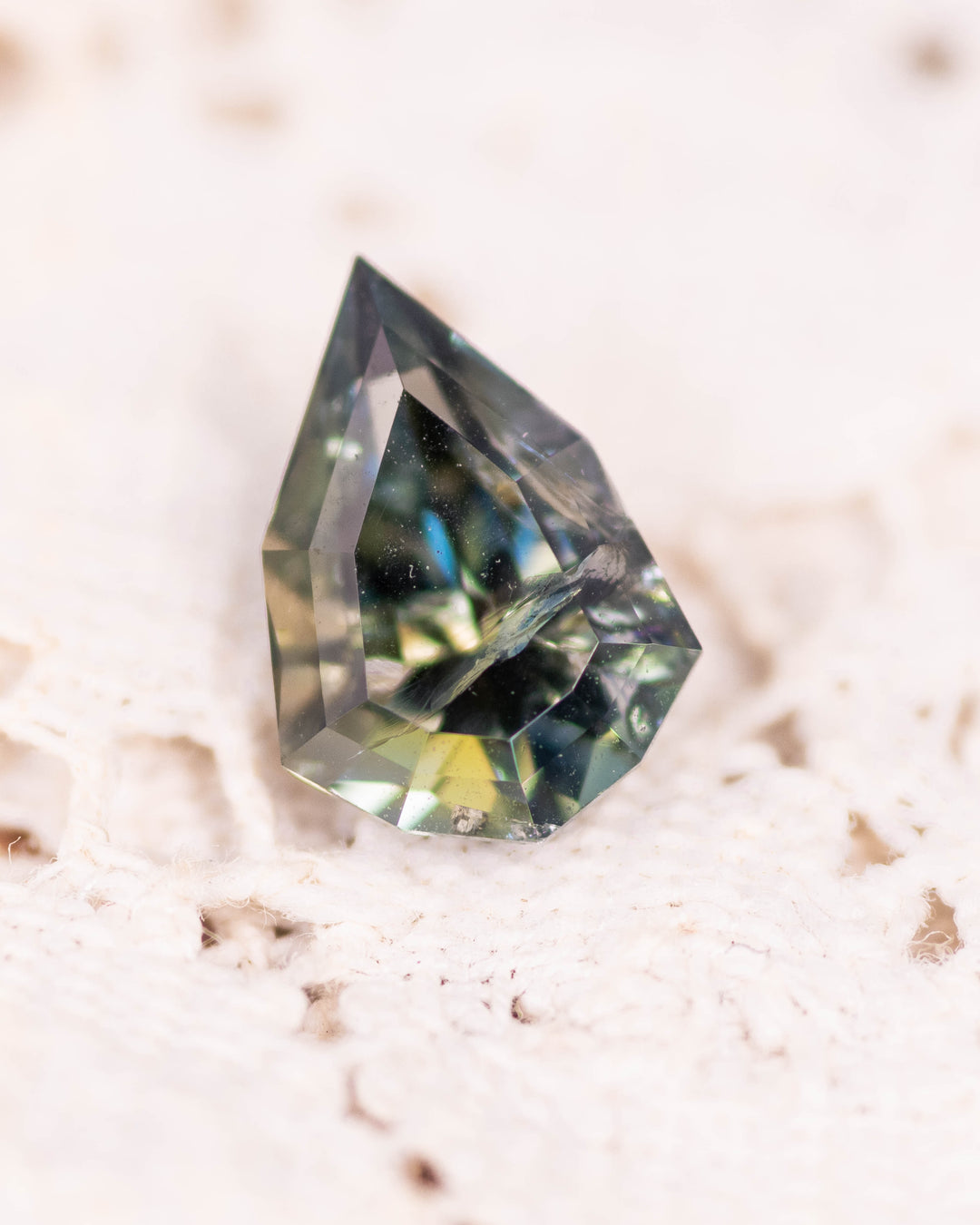 Macro image of 1.64CT yellow green teal tanzanite resting on lace fabric, highlighting color transitions and geometric symmetry of the custom mixed cut shield design, unheated gemstone from Merelani Hills, Tanzania.