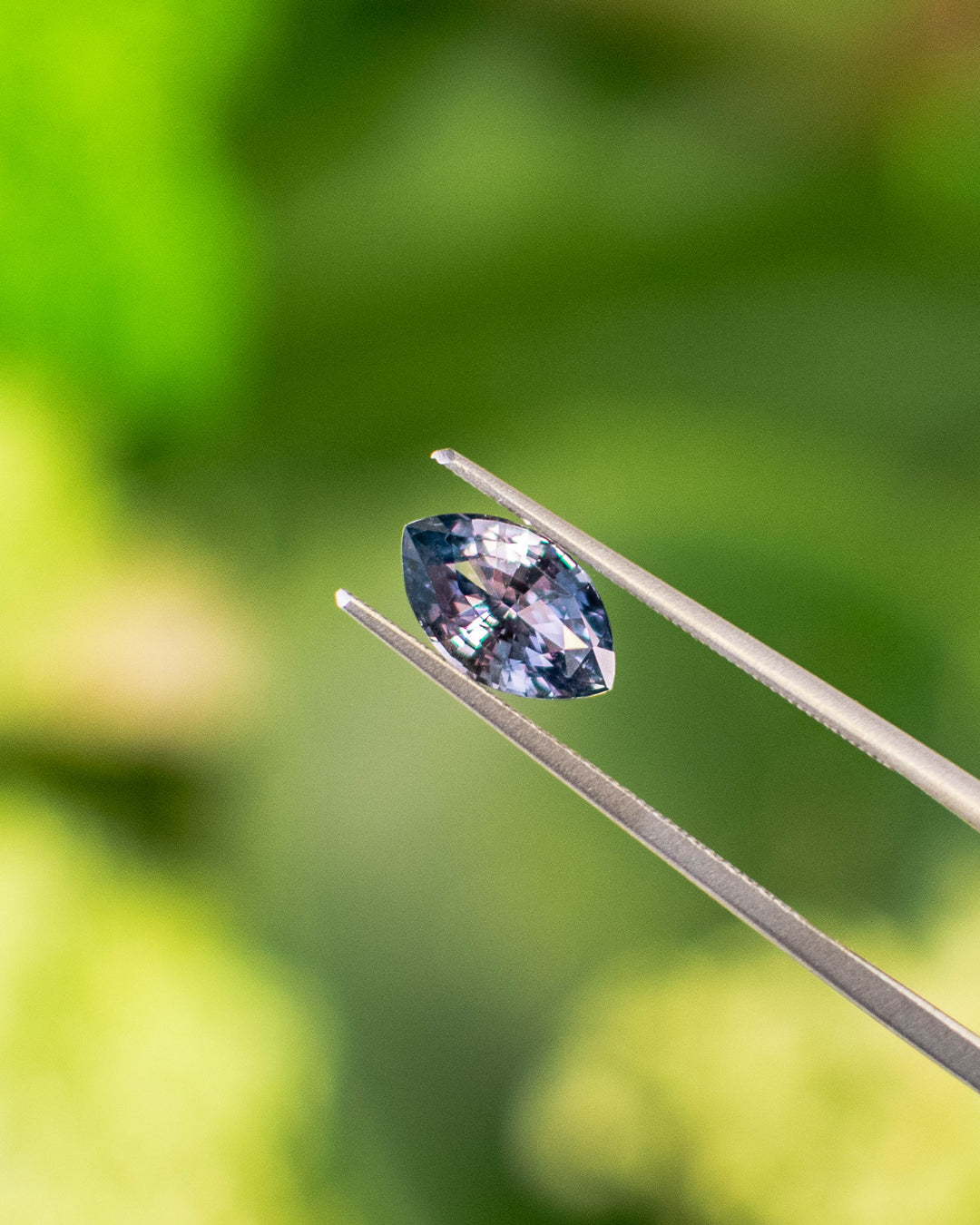 Close-up of 1.68CT color-shift sapphire held in gemstone tweezers under natural light, highlighting reflective facets and transitions between violet and gray tones in the precision marquise mixed cut, unheated sapphire from Madagascar.