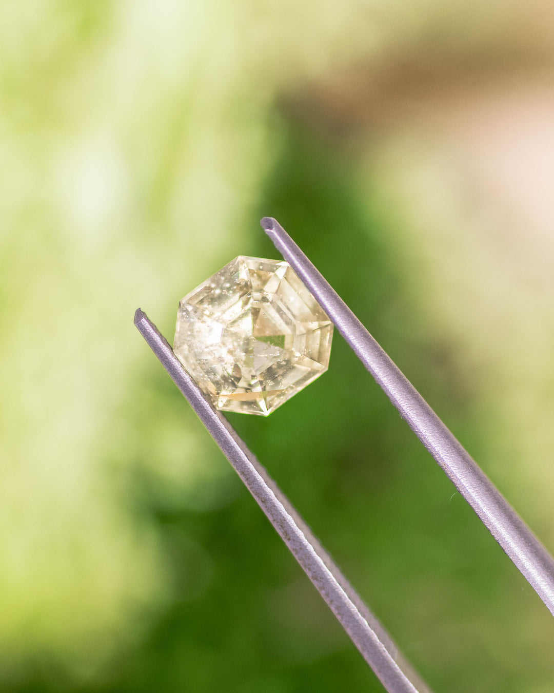 1.88CT teal yellow sapphire held delicately in gemstone tweezers with green natural background, capturing step-cut symmetry and sparkle of this unheated Asscher cut sapphire from Umba River Valley, Tanzania.