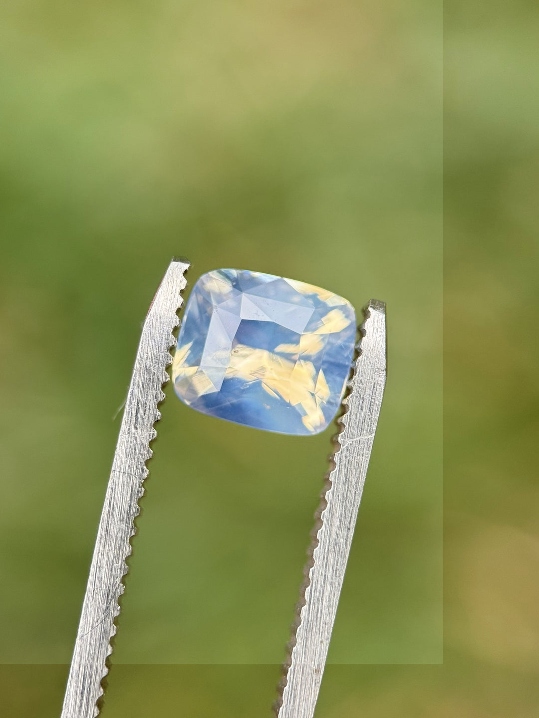 Natural 2.00CT sapphire with silver-blue opalescent tones held by tweezers against blurred green background, highlighting squared cushion faceting and soft internal reflections, unheated gem from Sri Lanka.