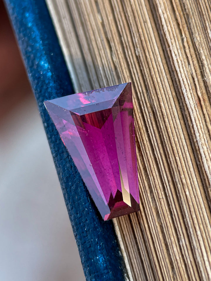 Natural tapered baguette garnet gemstone, 2.02 carats, resting elegantly on a textured surface, highlighting its radiant red and purple tones.