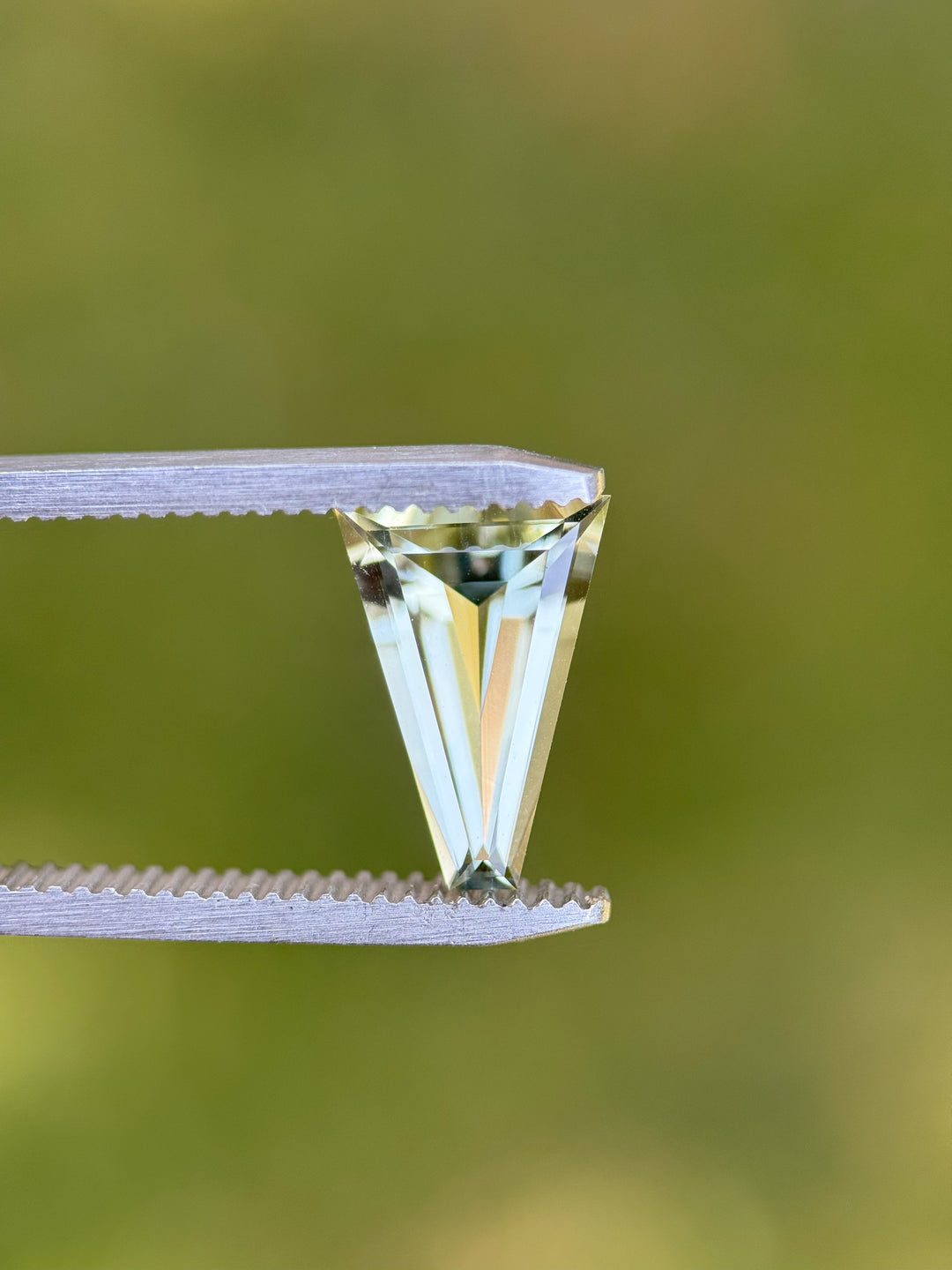 Close-up of mint green 2.02CT tourmaline held in gemstone tweezers against natural green background, displaying sharp custom tapered baguette step cut and clean clarity, no-heat gem from Congo.