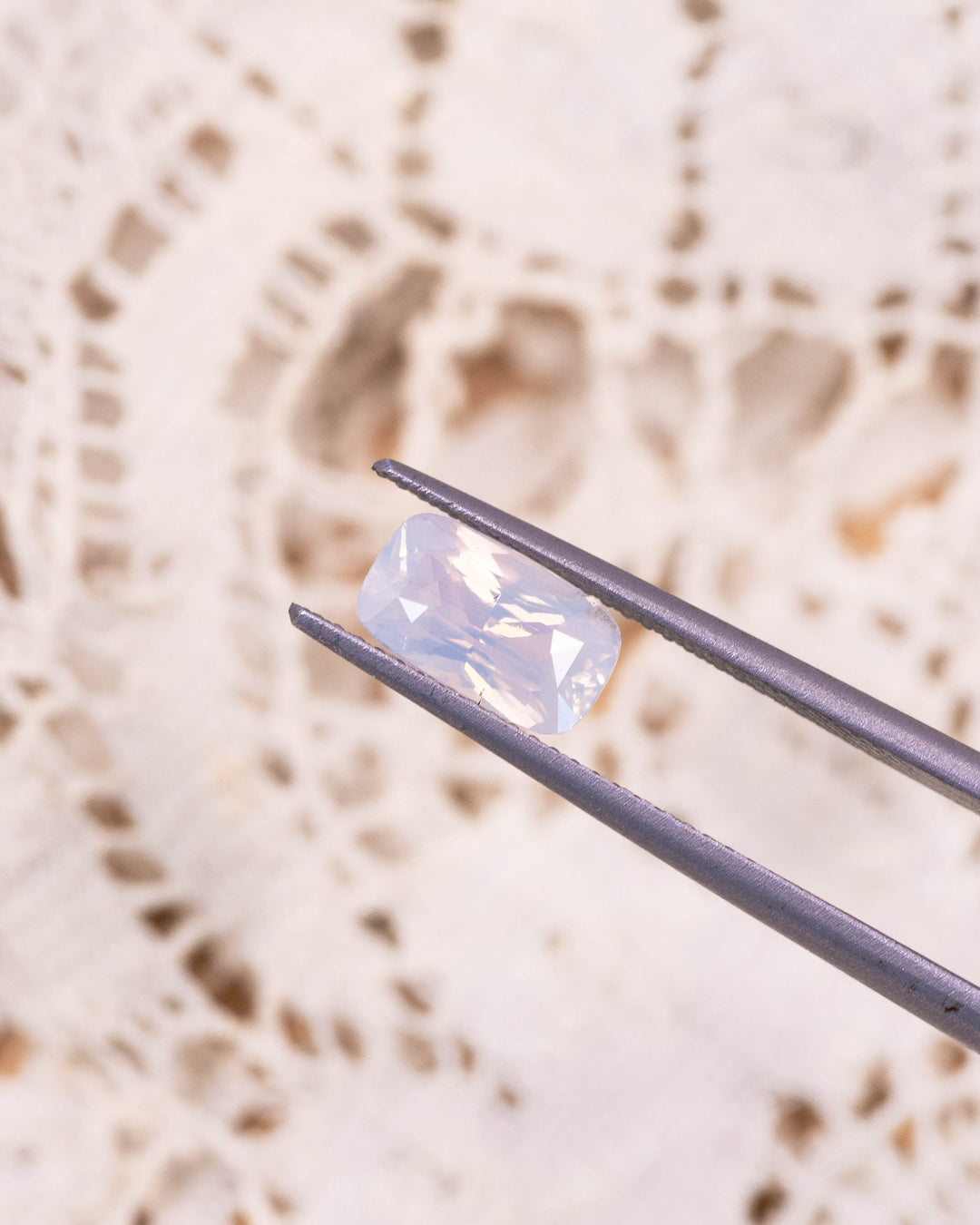 Macro image of 2.05CT opalescent peach sapphire held in gemstone tweezers, highlighting the soft peach glow, clean facets, and brilliance of the cushion cut, unheated sapphire from Madagascar.