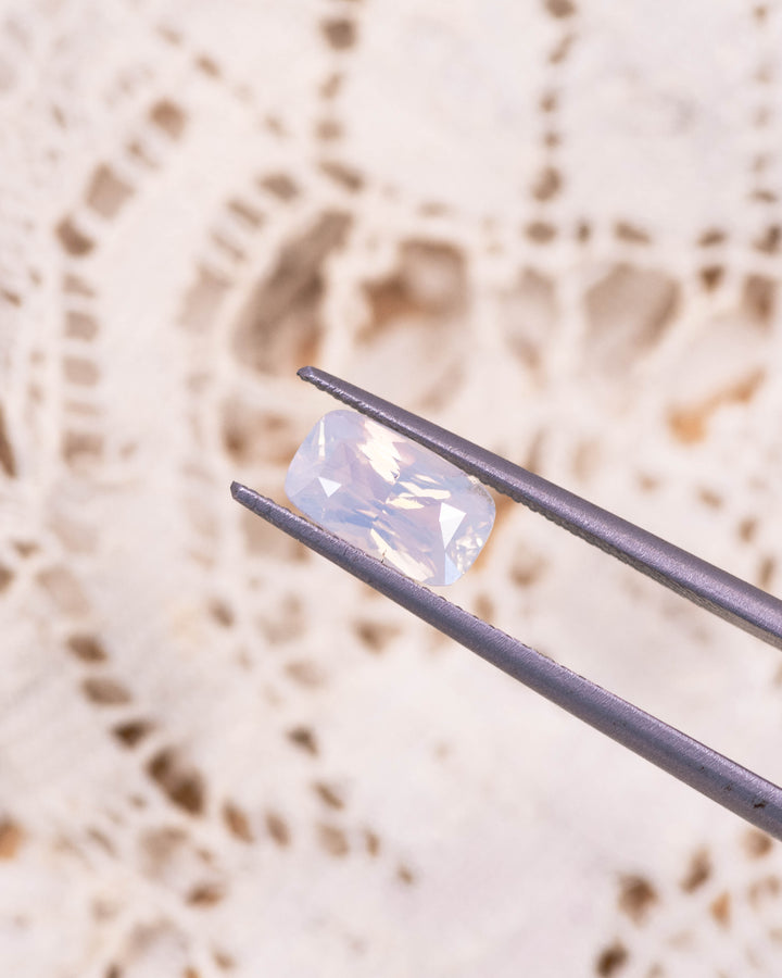 Macro image of 2.05CT opalescent peach sapphire held in gemstone tweezers, highlighting the soft peach glow, clean facets, and brilliance of the cushion cut, unheated sapphire from Madagascar.