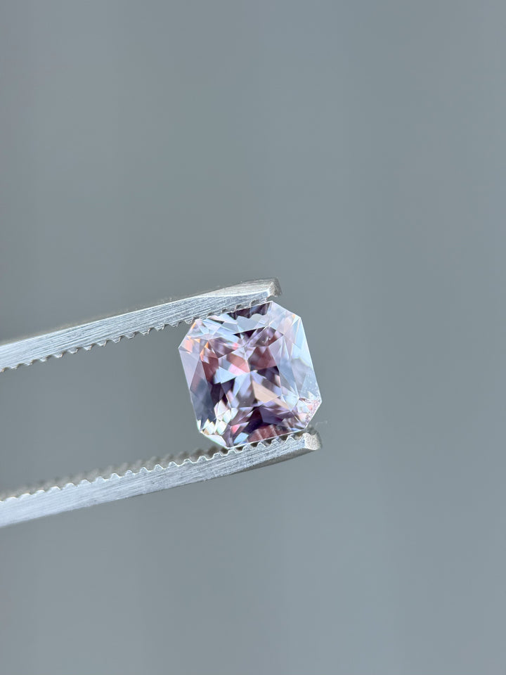 A pastel purple and pink 2.14-carat sapphire held in fine-toothed gemstone tweezers, captured against a smooth light gray background, highlighting the sparkle of the custom radiant cut.