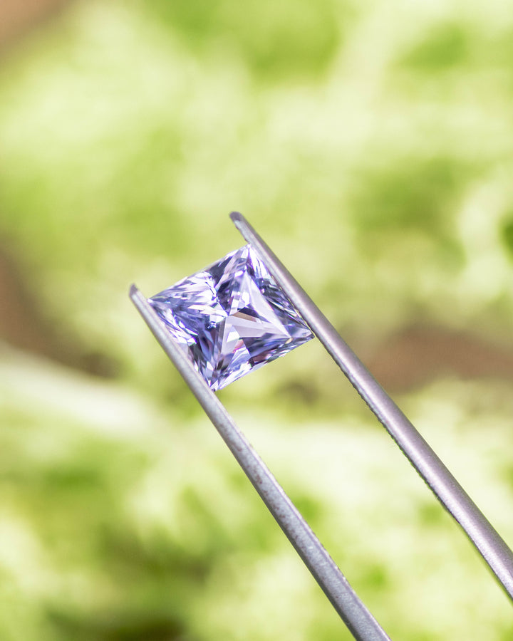 Macro shot of 2.15CT sapphire held in gemstone tweezers with a natural green background, highlighting gray and lavender hues and sharp facets of the custom kite cut, unheated sapphire from Sakaraha, Madagascar.