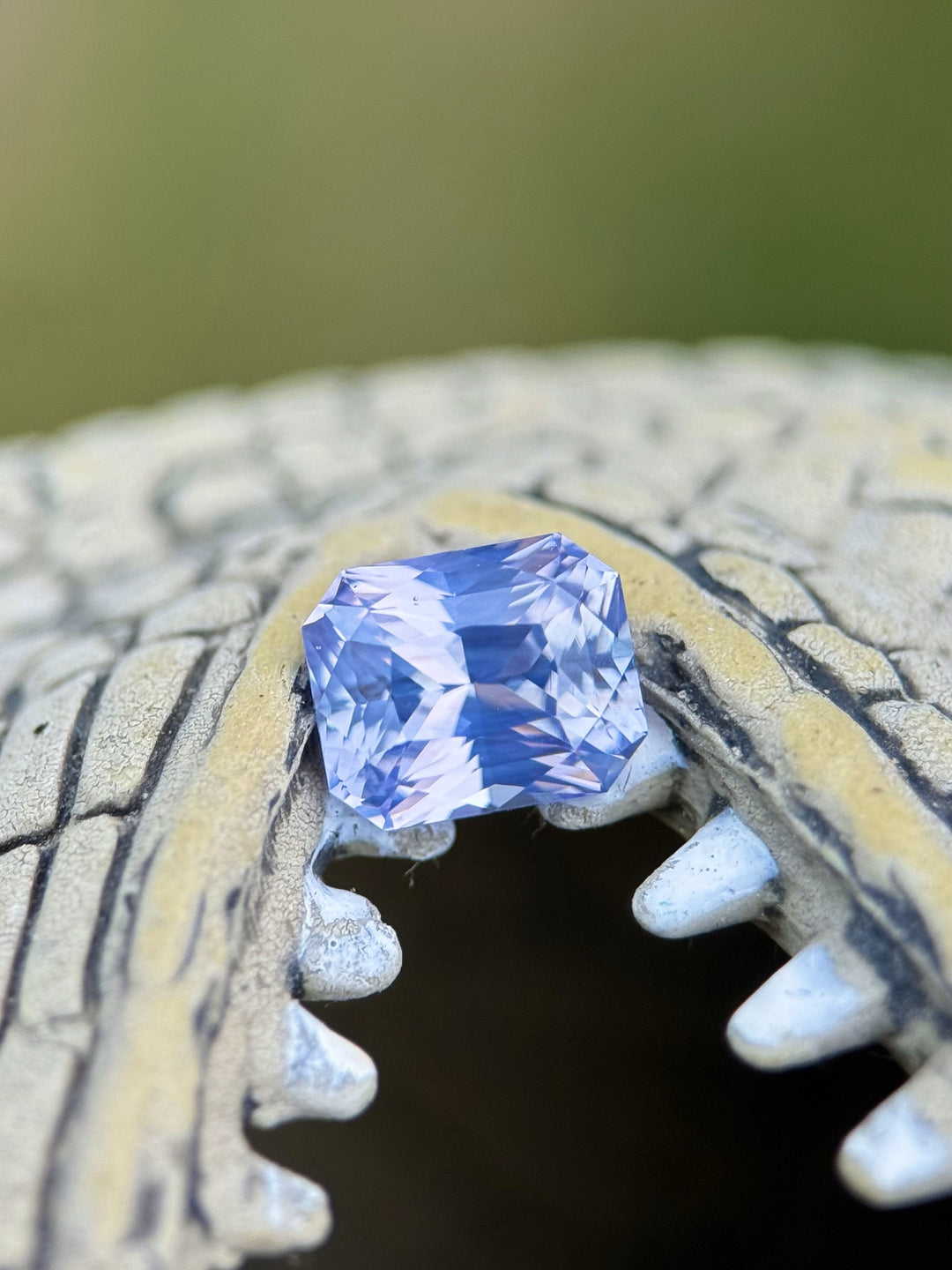 Macro image of a 2.16ct unheated sapphire placed on a natural textured surface, showcasing its radiant cut and unique pastel color blend of peach, lavender, and violet.
