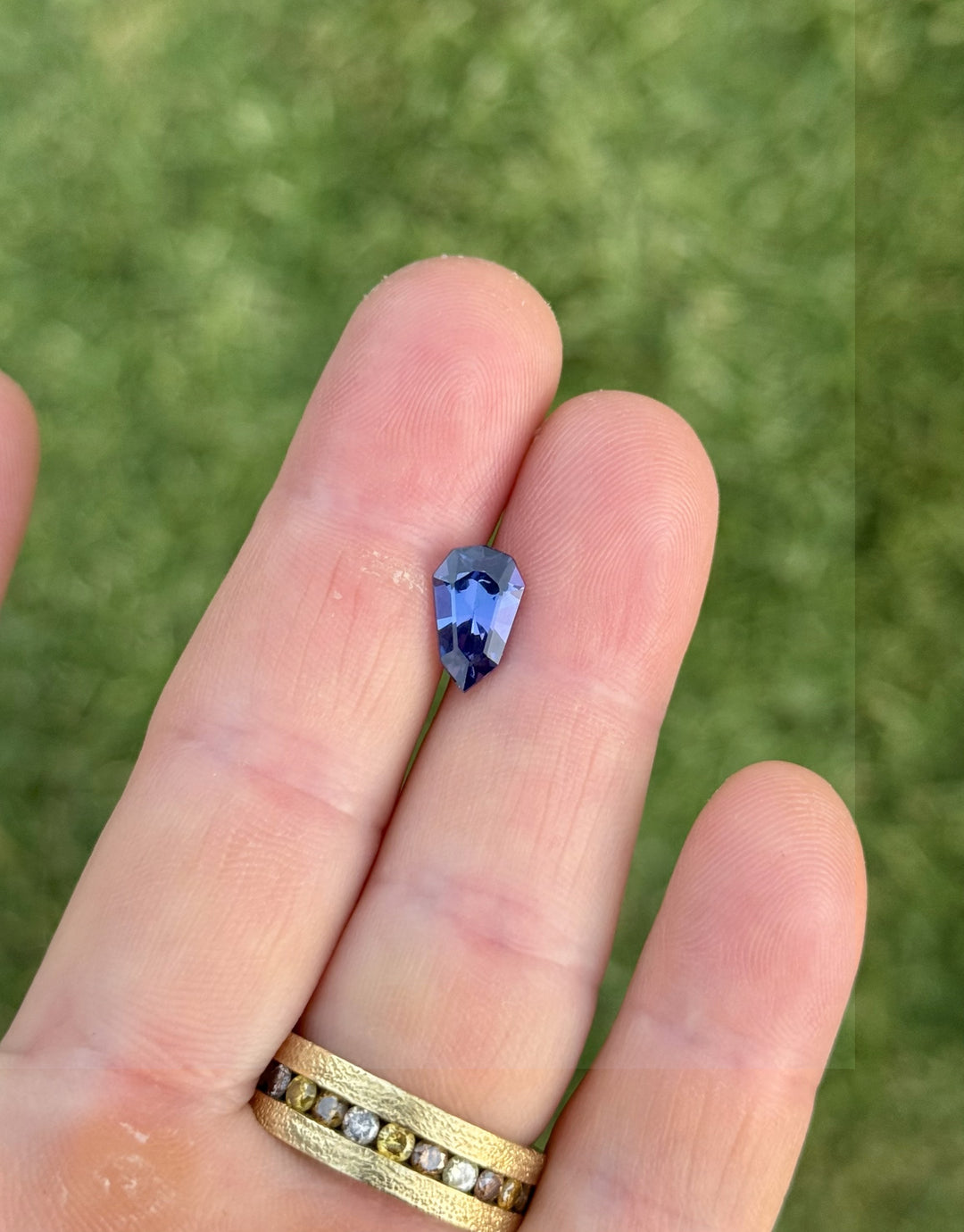 Close-up photo of 2.42CT custom elongated shield cut tanzanite held between fingers under natural sunlight, showing deep blue and purple tones with flashes of mermaid green, unheated gem from Tanzania.