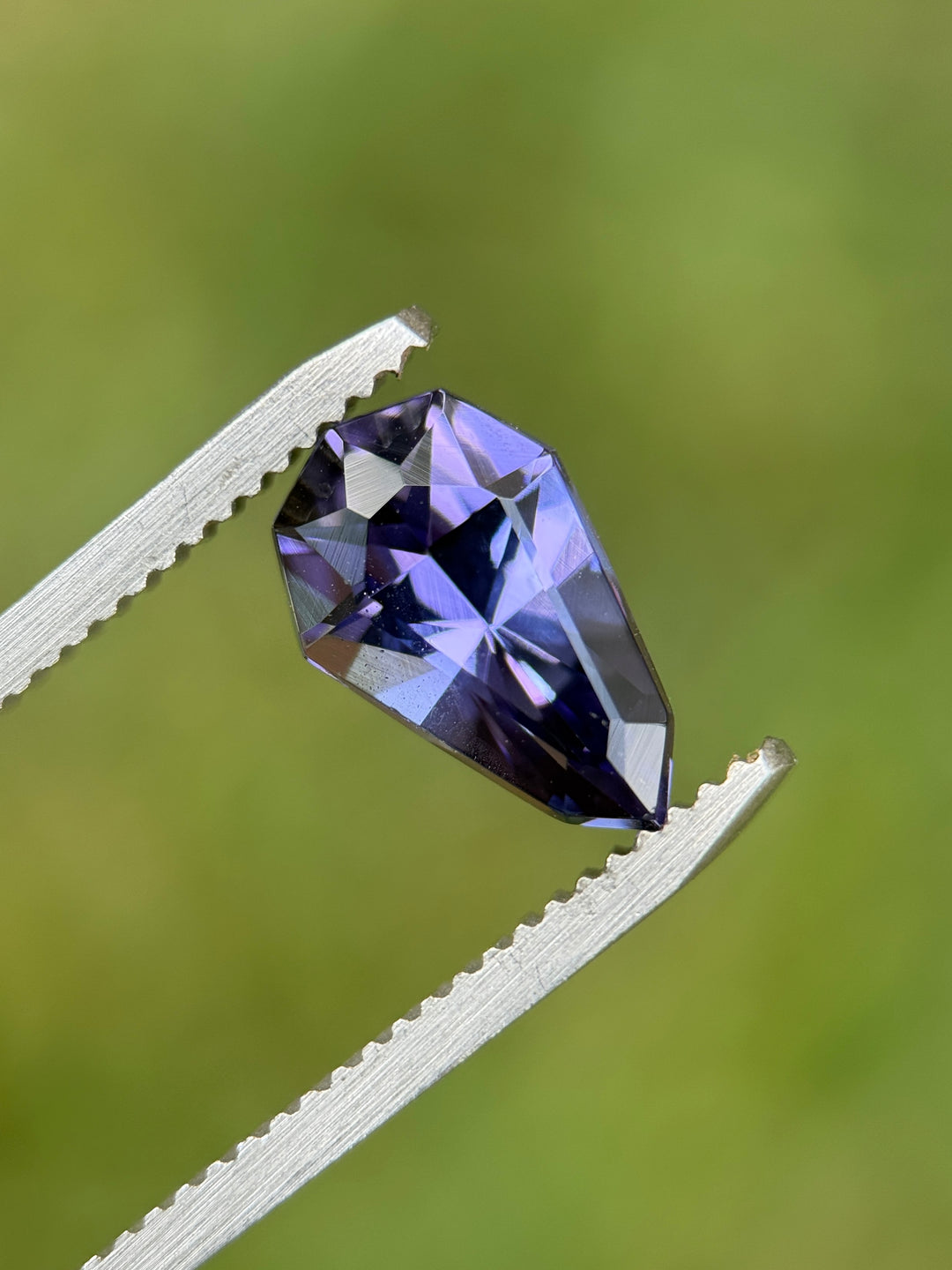 Close-up of 2.47CT Tanzanite gemstone held in tweezers against a green background, highlighting slightly greenish blue and violet mermaid hues with precise shield mixed cut facets, natural and untreated from Tanzania.