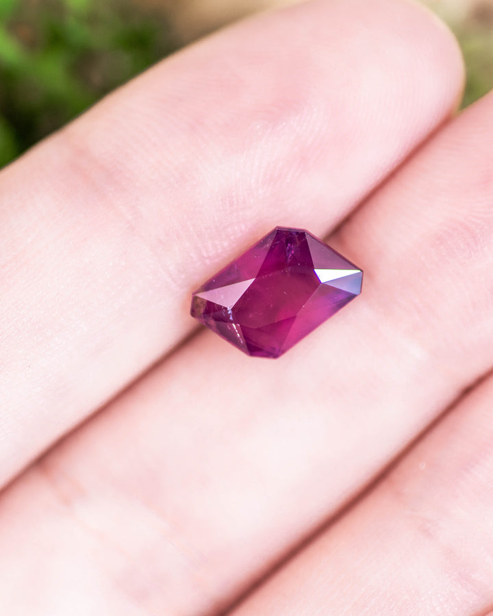 2.85 carat sapphire with vivid pink, purple, and red hues resting on fingertip, displaying geometric radiant emerald cut facets and brilliance of this untreated gemstone from Umba River Valley, Tanga, Tanzania.