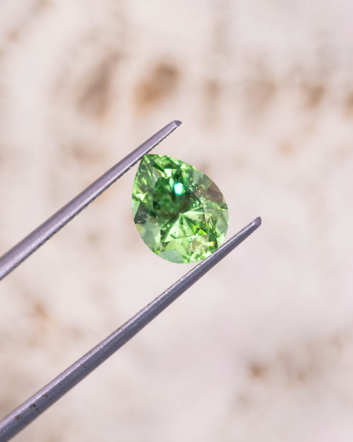 Close-up photo of 2.85CT lime green Tsavorite garnet held in gemstone tweezers under natural light, highlighting sparkle, clarity, and faceting precision of the modified brilliant pear cut, unheated Tsavorite from Kenya.