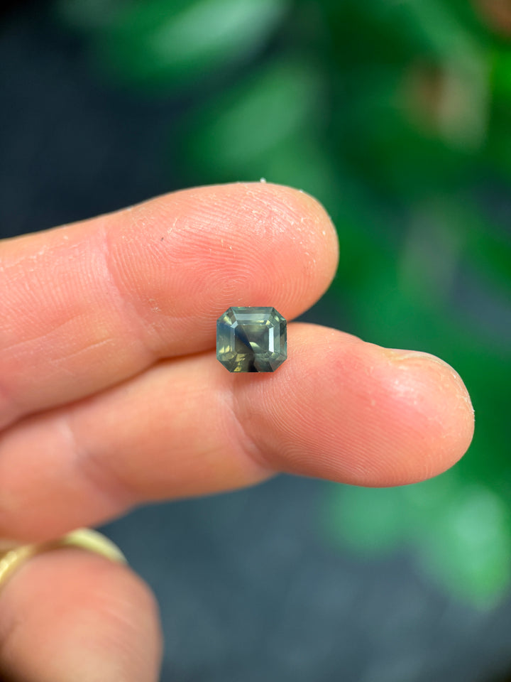 3.40 carat natural sapphire balanced on fingertip, emerald cut opalescent sapphire with earthy olive, teal, brown, and blue hues from Nigeria