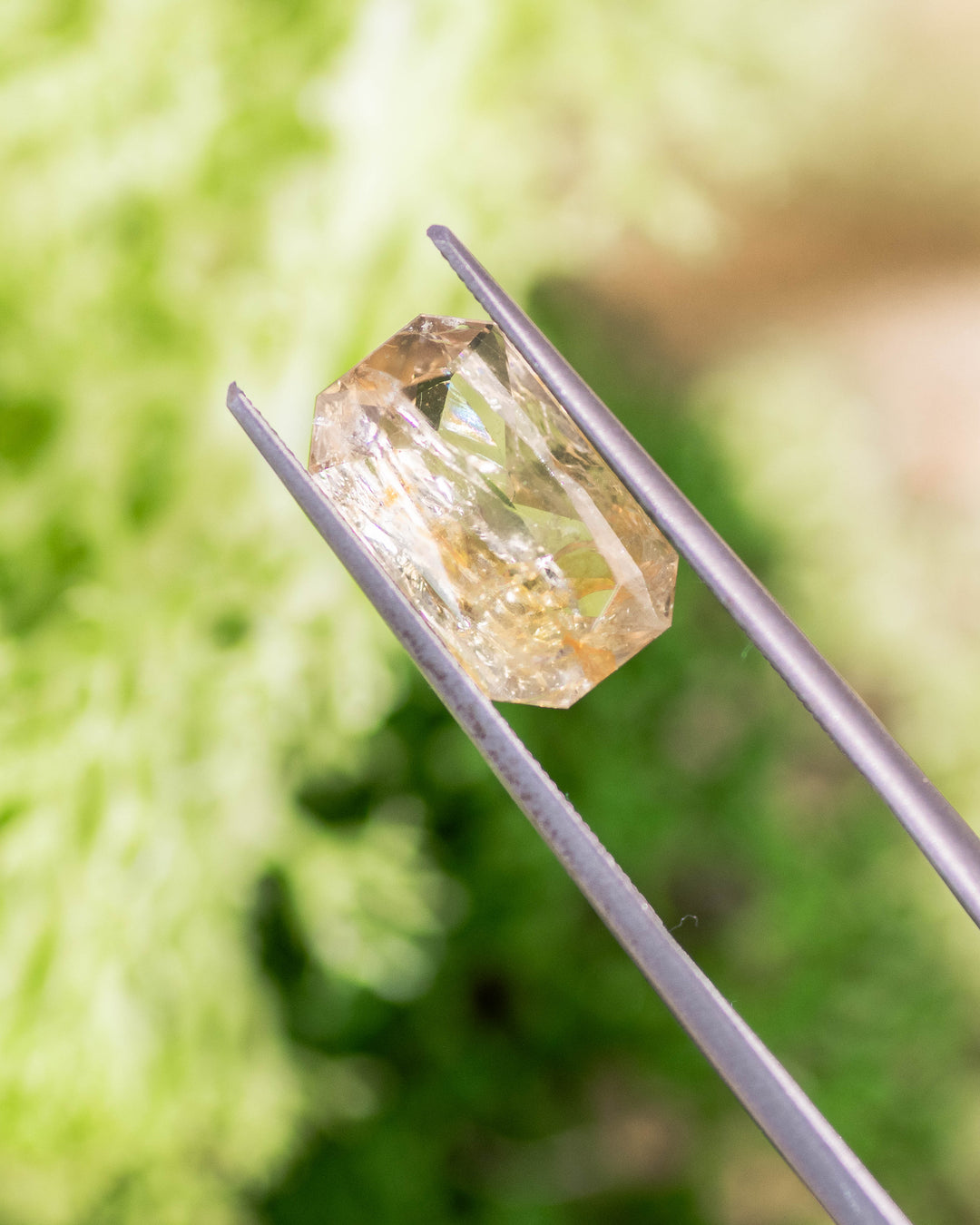 Macro photo of 3.50CT yellow and orange sapphire held in gemstone tweezers outdoors, capturing internal clarity and brilliance of its custom modified radiant cut, unheated sapphire from Umba River Valley, Tanzania.