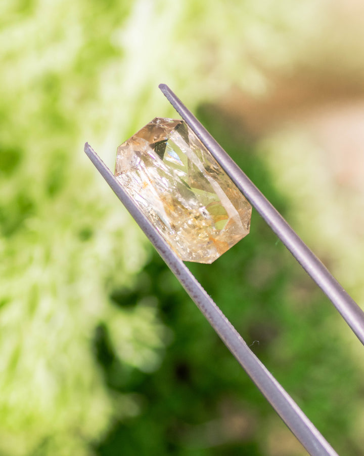 Macro photo of 3.50CT yellow and orange sapphire held in gemstone tweezers outdoors, capturing internal clarity and brilliance of its custom modified radiant cut, unheated sapphire from Umba River Valley, Tanzania.