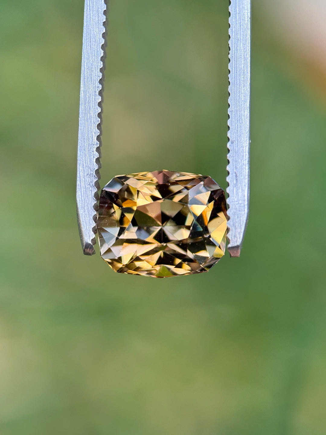 Close-up of 3.81CT Tanzanite held in gemstone tweezers against a green background, showing a vibrant mix of lavender pink and golden yellow hues in a precision geometric cushion modified brilliant cut, untreated gem from Merelani Hills, Tanzania.