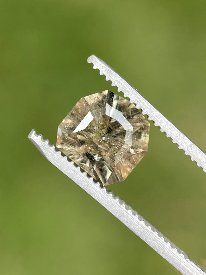 Close-up of a custom calf head cut 3.82ct sapphire in tweezers, displaying grey, pink, and peach tones.