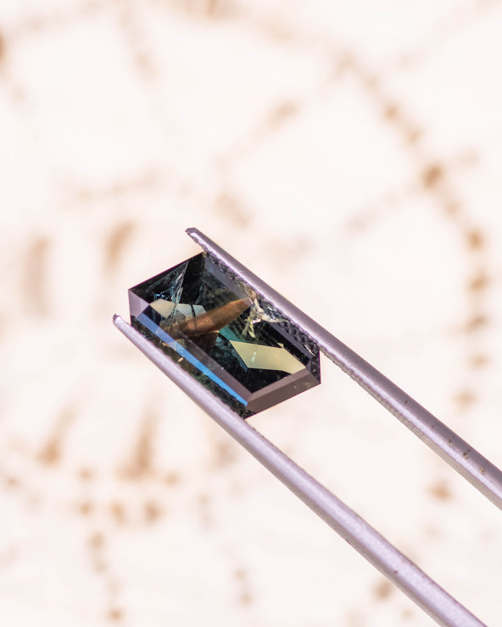 3.93CT peacock teal sapphire held delicately in gemstone tweezers against a neutral background, capturing its deep oceanic tones and geometric symmetry of the custom rhombus mixed cut, unheated sapphire from Garba Tula, Kenya.