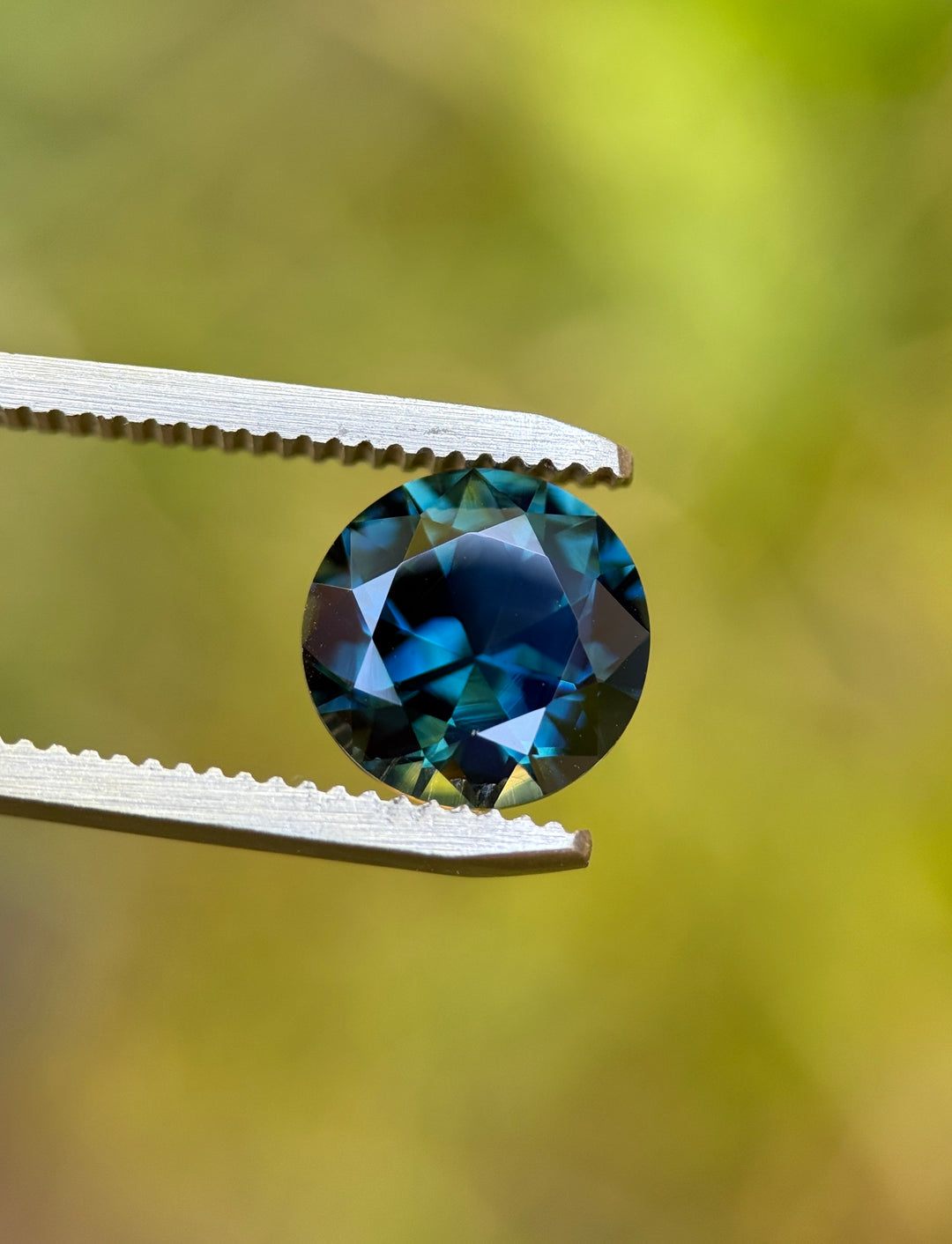 Round brilliant cut 3.98ct peacock teal blue green sapphire held in gem tweezers, natural untreated Nigerian sapphire displaying precision cut and vivid color