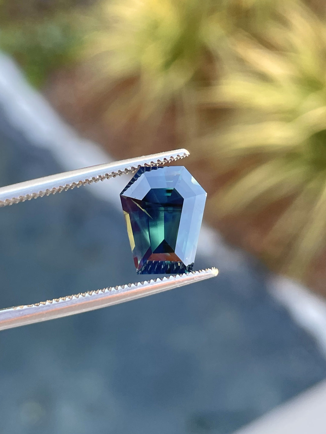 A stunning 4.65-carat sapphire with forest green, teal, and yellow hues in a step cut coffin shape, resting on a textured natural surface.