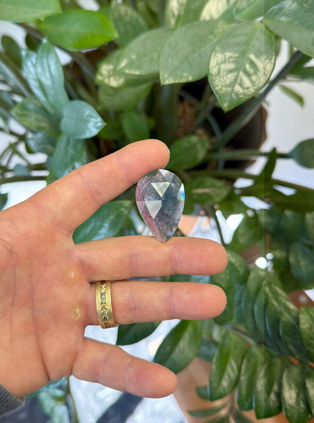 Large 46.80CT pear rose cut tourmaline held between fingers in front of green leaves, displaying its natural purple, pink, and blue coloration under daylight, mined in Paprok, Afghanistan.