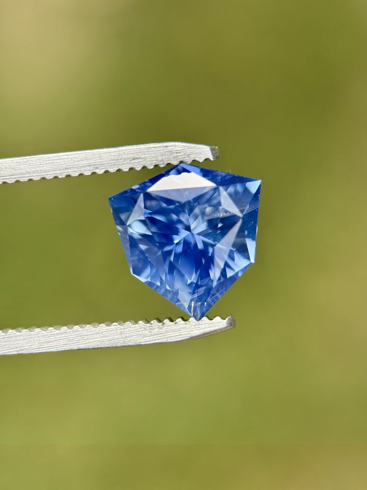 Close-up of 5.10CT bright blue sapphire held in gemstone tweezers, highlighting the stunning symmetry, facet alignment, and vivid saturation of the precision custom trillion shield mix cut, unheated sapphire from Madagascar.